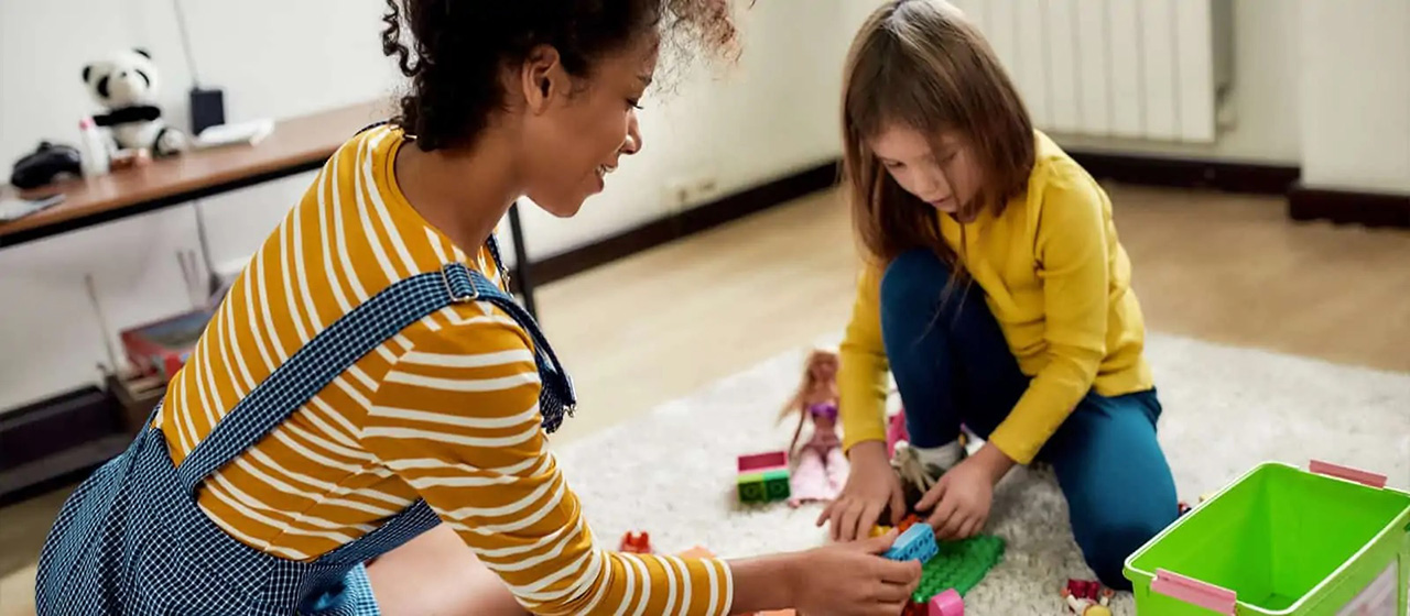 A female babysitter plays with a young girl using lego blocks