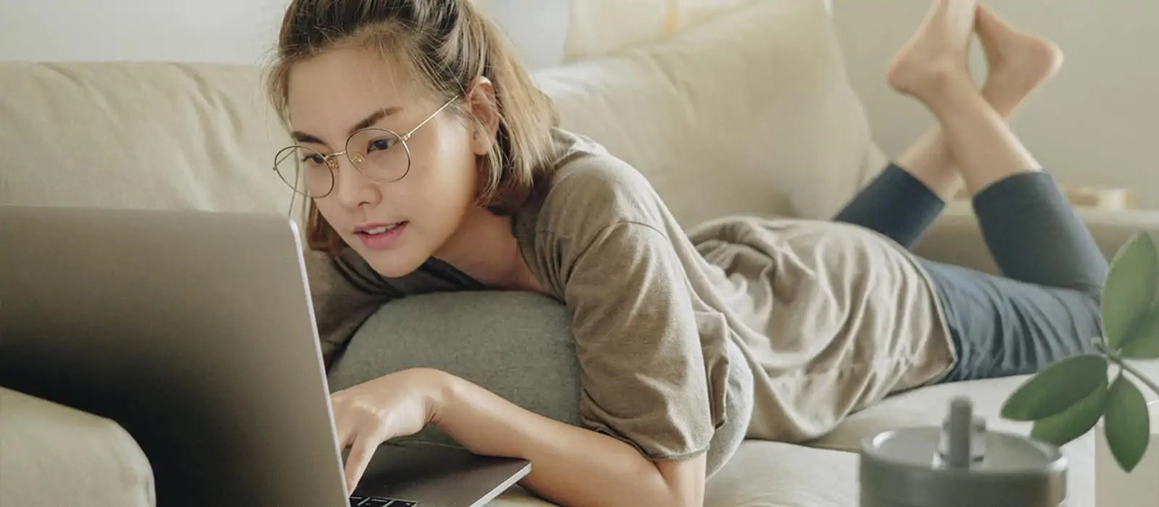 Female with glasses laying on a couch on her laptop with her feet crossed behind her