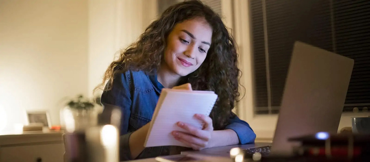 Female writing in a notebook with her laptop open in front of her